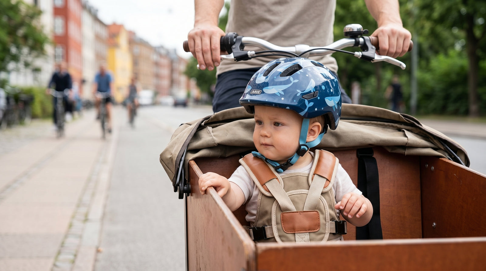 Baby med blå Abus Smiley cykelhjelm fastspændt i ladcykel — dansk by, far cykler